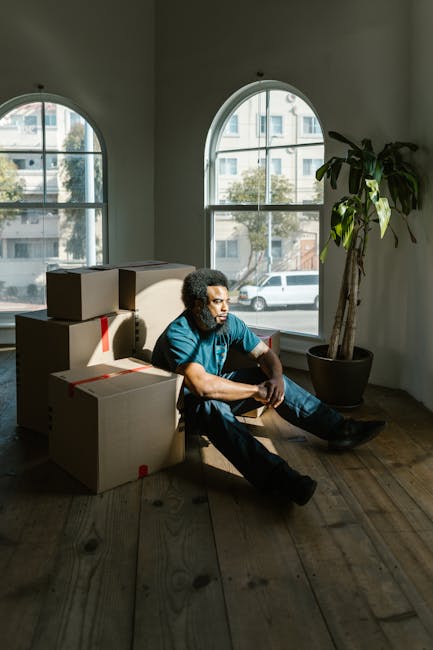 A man with curly hair and a beard, wearing a blue t-shirt and dark trousers, is sitting on the floor inside a room with wooden flooring and large arched windows that let in natural light. He is surrounded by several cardboard boxes, some sealed with red packing tape, indicating they are packed for a house move. The boxes are stacked to his left and behind him, suggesting he is in the process of packing or unpacking during a house relocation. The room contains a tall potted plant with broad green leaves inside a black pot positioned next to the window on his right. Outside the windows, a street scene with parked cars and multi-storey buildings is visible, hinting at an urban setting. This scene likely depicts a break during a furniture transport or home moving process, illustrating elements of packing, loading, and logistics typical of a professional removals service by [COMPANY_NAME]. The overall environment and scene are professionally staged with natural lighting, emphasizing the interior space involved in house removals and packing and moving activities.