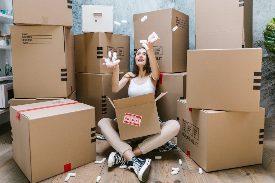 A young woman with light skin and long dark hair is sitting cross-legged on a wooden floor inside a room, surrounded by multiple large cardboard boxes sealed with red and black tape. She is wearing a white sleeveless top, beige pants, and black-and-white sneakers. The woman is smiling and tossing white packing peanuts into the air, some of which are falling into an open cardboard box in her lap that has a red 'Fragile' label on it. The boxes vary in size and are stacked or positioned around her, with a few placed directly on the floor and others arranged against a wall with a light blue textured finish. In the background, a small white microwave is visible on a shelf, and a potted plant with green leaves sits on the side. The scene depicts a home packing or moving process, with attention to careful handling and organization typical of house removals and furniture transport services offered by Man with Van Grange Park.