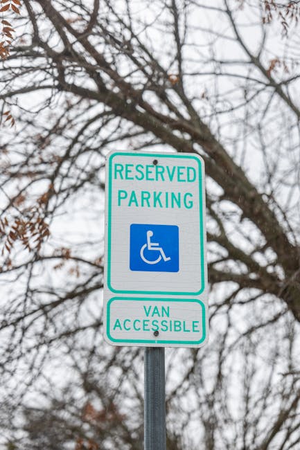 A vertical metal signpost with a green and white reserved parking sign indicating a designated space for wheelchair users and van accessibility, featuring a blue wheelchair symbol. The sign is mounted outdoors against a background of leafless tree branches and an overcast sky, suggesting a parking area adjacent to a residential property in Grange Park. The context reflects urban parking regulations relevant to house removals and moving logistics, with the sign positioned near a property entrance or public pavement, as seen in the service offered by Man with Van Grange Park for home relocation and furniture transport services.