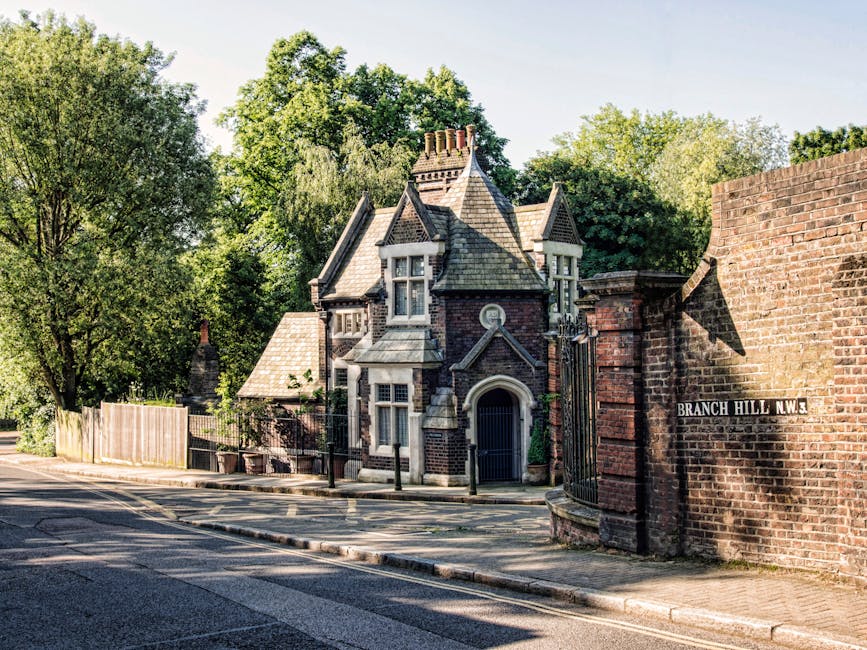 A Victorian-style house situated on a tree-lined street in Grange Park, with a grey brick façade, ornate window frames, and a peaked roof. The property is enclosed by a low brick wall with an iron gate, partially visible behind a wooden fence. The street is paved with asphalt and features a curb with a sidewalk, where a black lamppost and a street sign reading 'Branch Hill N.W. 3' are mounted on a brick wall to the right. Surrounding the house are lush green trees casting shadows onto the pavement, indicating daylight. The scene depicts a quiet residential area suitable for home relocation or furniture transport services, as provided by Man with Van Grange Park, with the house awaiting potential removal or moving process involving loading and transport preparations.