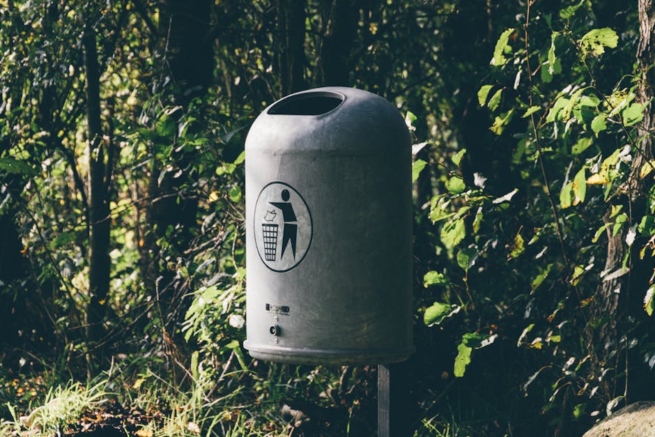 A metal waste bin with a rounded top and an open slot for disposing of trash, positioned outdoors in front of dense green foliage with various leaves and branches. The bin features a black and white pictogram on its front, depicting a person tossing rubbish into a bin, indicating its purpose for waste disposal. The bin is mounted on a metal post and is surrounded by grass and small plants at the base. This scene relates to the process of clearing out household items during a home relocation or furniture transport, relevant to house removals and packing and moving services offered by Man with Van Grange Park. The natural lighting suggests daytime, with sunlight filtering through the leaves, highlighting the outdoor environment typically encountered during the disposal of bulky waste before move logistics.