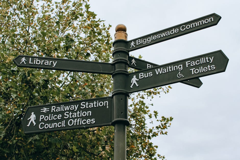 A black metal directional signpost outdoors against a backdrop of green and orange-leaved trees, displaying multiple white directional signs with icons and text. The signs point towards various locati