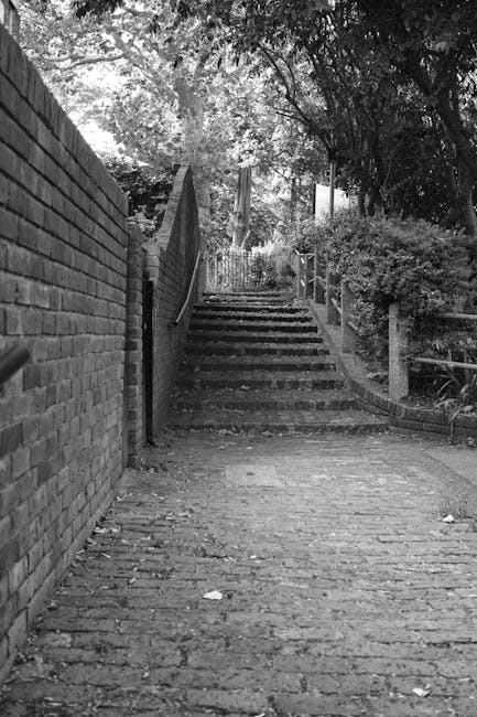 Two movers from Man with Van Grange Park are ascending a set of outdoor stairs at a railway station or transit area, both carrying large black backpacks and holding a piece of furniture, possibly a sofa or similar item, wrapped in plastic or fabric to protect it during the loading process. The stairs are made of dark material with a metal handrail on one side, leading up to a covered platform with a modern, open structure featuring metal pillars and a roof. The environment is well-lit with natural daylight, and the movers appear to be engaged in a home relocation or furniture transport task as part of a house removals service, focusing on safely navigating the stairs while moving bulky items. The scene reflects a typical moving logistics phase within a packing and loading process, supported by the professional team at Man with Van Grange Park.