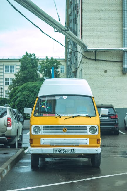 A yellow and white Volkswagen van parked in a designated parking area near a commercial building, with several other vehicles visible on either side. The van is positioned facing the camera, showing its front grille, windshield, and headlights, with a few decorative items hanging from the interior rearview mirror. The surrounding environment includes a brick building on the right, overhead electrical wires, and green trees in the background. The parking space appears to be used for home relocation or furniture transport, as part of the loading process managed by Man with Van Grange Park. The scene is outdoors during daylight, with natural lighting illuminating the vehicles and nearby pavement, indicating an area suitable for van-based removals and moving services near Grange Park Station.