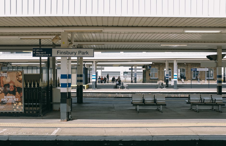A view of Finsbury Park railway station platform showing an overhead sign with the station name, surrounded by a metallic roof structure. Several passengers are seated on metal benches located on the platform, with some standing and walking nearby. In the foreground, there are two empty wooden benches and a small advertisement board on the left side displaying a poster for 'Romeo and Juliet.' The platform surface is paved with concrete, featuring tactile paving strips near the edge for accessibility. Visible elements include a black metal fence, a ticket machine, and a track separating the platform from the approaching train area. The environment suggests an urban setting during daylight, with natural lighting illuminating the scene. This context aligns with home relocation and furniture transport logistics by highlighting the importance of parking and loading areas near railway stations, which can be relevant for planning moves or transport services for customers of Man with Van Grange Park.