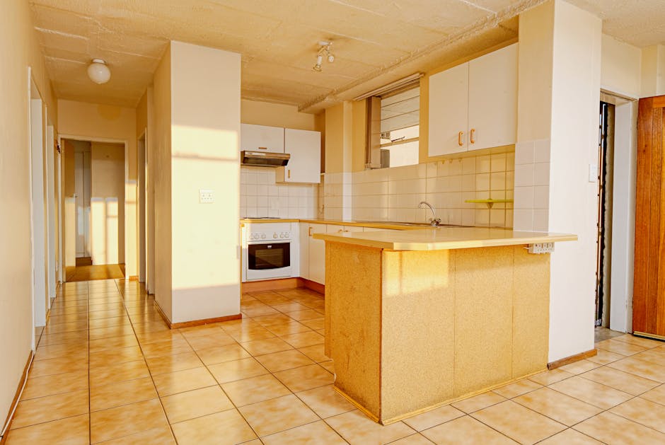Interior view of a kitchen during a home relocation, showing a tiled floor and cream-colored walls illuminated by natural sunlight. The kitchen features white cabinets with orange handles, a white oven with a built-in stovetop, and a stainless steel sink beneath a window. A small yellow shelf is mounted on the wall above the sink. In the foreground, there is a yellowish kitchen island or breakfast bar with a tiled surface, positioned near the perimeter of the room. To the right, a doorway reveals a corridor leading to other parts of the house, while a wooden wardrobe is visible in the background. The area appears tidy, with no visible packing materials or boxes, suggesting a staged or unprepared space for moving. Man with Van Grange Park is familiar with such interior environments, facilitating efficient packing and furniture transport during house removals or relocation services at this property.