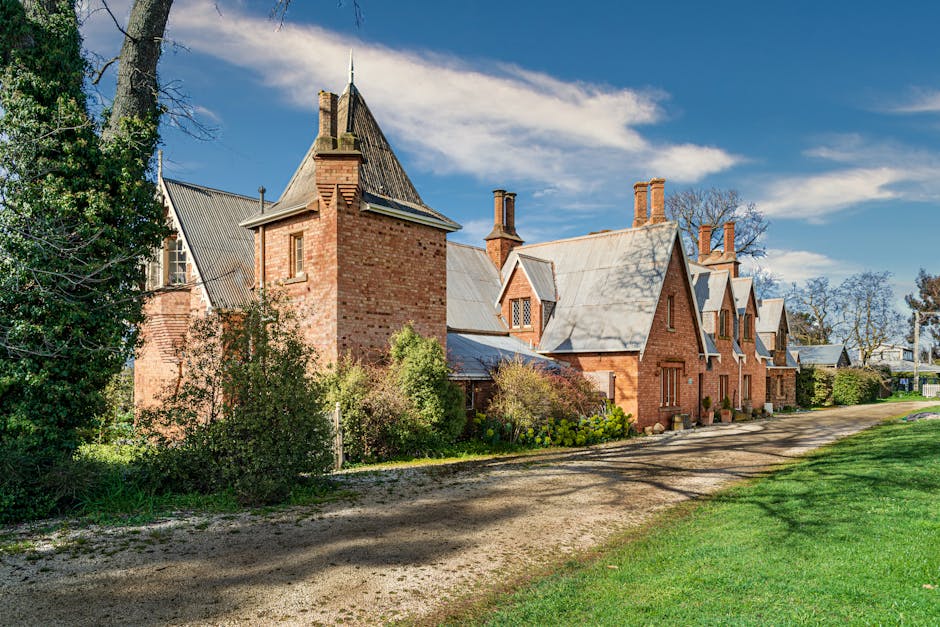 A large, red brick country house with a steeply pitched grey slate roof, multiple chimneys, and small dormer windows under a bright blue sky with wispy clouds. The house features a tower-like section with a pointed roof and a chimney, surrounded by well-maintained garden bushes and trees. A gravel driveway runs alongside the building, with patches of green grass on either side. The scene is set during daytime with natural sunlight illuminating the house exterior, suggesting a setting suitable for home relocation or furniture transport activities. The image captures the entire facade of the house and part of the adjacent garden and driveway, providing a clear view of the property’s structure and environment, reflecting the type of property typically involved in house removals or moving services.